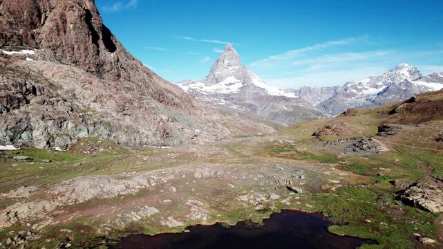 Cinematic aerial reveal of glacial lake reflection of the iconic Matterhorn towering over the Zermatt valley. Pristine alpine scenery featuring rocky terrain, and clear blue skies in the Swiss Alps.