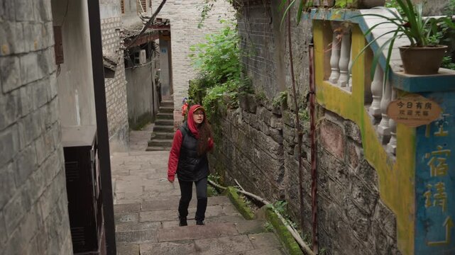 Asian woman exploring colorful heritage alley past painted pillar and potted plants, wearing red jacket and cap, upbeat stride on stone steps, lively textures and script signs, tourist discovery