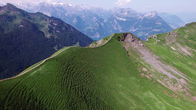 Panoramic alpine ridge covered in green grass, winding mountain trail leading toward distant peaks and a turquoise lake, captured on a clear summer day by 4k aerial drone