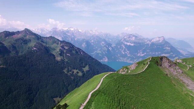 Drone aerial of hikers along Stoos Ridge in the Swiss Alps, overlooking Lake Lucerne, with lush green slopes and snow-capped peaks under a clear summer sky.