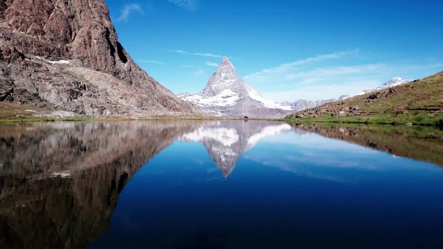 4K drone shot of the Matterhorn reflected perfectly in a calm alpine lake, surrounded by rocky terrain and clear blue skies, creating a serene and iconic Swiss mountain landscape.