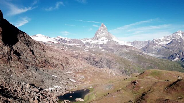 4K drone aerial of the iconic Matterhorn rising above rocky alpine terrain and distant snowy peaks, showcasing Switzerland&rsquo;s dramatic mountain landscape under clear blue skies.
