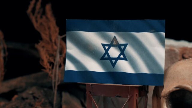 Conceptual scene of an israeli flag waving over a diorama with toy soldiers, a skull, and a lantern, representing conflict and remembrance