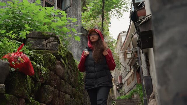 Asian woman walking mossy stone steps, red hoodie black vest, backpack, admiring hanging red flowers on weathered stone wall, green moss and foliage, narrow village lane, calm reflective mood,