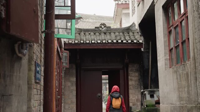 Backpacker walking through historic narrow alley past weathered walls and tiled roof, orange backpack and red jacket, approaching ornate wooden gate, quiet cinematic mood, slow travel vibe, hidden