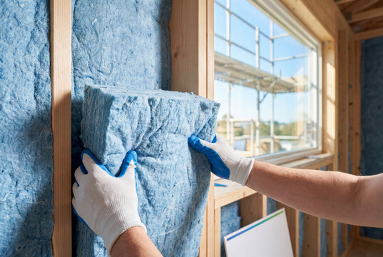 Construction worker installing blue thermal insulation batt into wooden wall frame