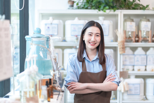 Proud and confident Asian shopkeeper in a brown apron stands with arms crossed and a bright smile inside her eco organic store, embodying female entrepreneurship and success.