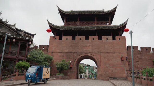 Overcast ancient gate with red lanterns street scene with blue delivery tricycle parked by brick arch flanked by traditional tiled roofs and stone walls muted colors cultural atmosphere quiet morning