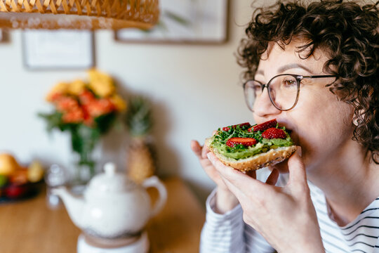 Middle aged woman taking bite of avocado toast with strawberries at home