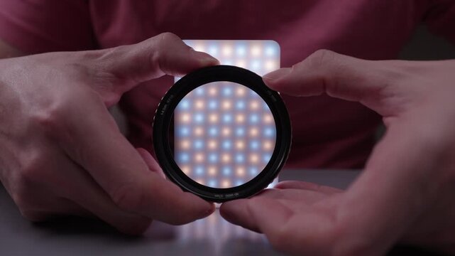 Photographer hands holding and rotating circular polarizing cpl filter in front of bicolor led light panel to demonstrate effect on light reflection and glare reduction in dark room, close-up, slowmo.