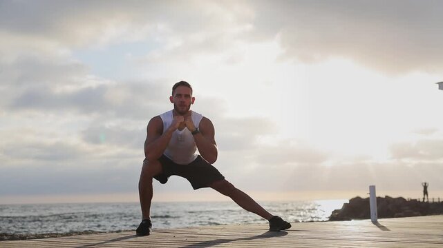 Full body young cool strong sporty athletic fit sportsman man wear sports clothes stretch legs do squats warm up train at sunrise sun dawn over sea beach outdoor on pier seaside in summer day morning