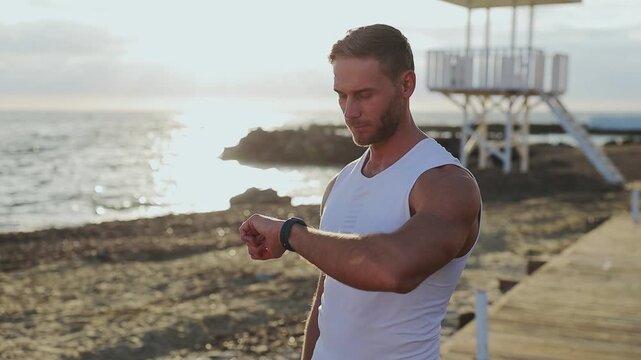 Young strong sporty athletic fit sportsman man wear sports clothes use mobile cell phone look at smart watch warm up train at sunrise sun over sea beach outdoor on pier seaside in summer day morning