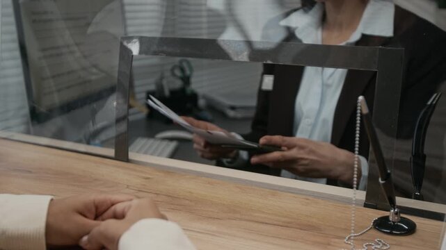 Over-shoulder medium close-up of female visa agency customer handing documents to smiling Caucasian clerk in formal suit, checking application form and asking questions on visit purpose