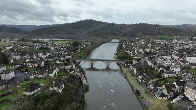 Aerial view of Argentat sur Dordogne with wide Dordogne river stone bridge and dense town buildings nestled between green hills under cloudy sky
