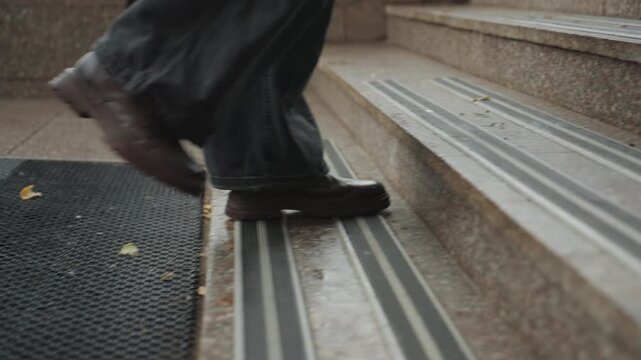 Closeup wet boots stepping mat, creator arriving on stone steps, rubber mat texture, slow deliberate pace, jeans hem brushing ground, rainy urban morning, tactile entrance detail