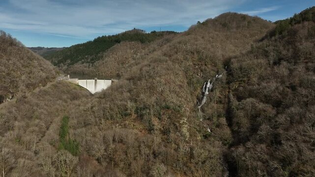 Aerial view of Hautefage dam in Correze nestled in a deep forested valley with a small waterfall visible on the hillside under blue sky