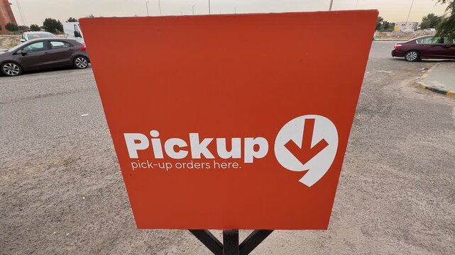 Bold orange pickup orders sign with white arrow logo displayed on a metal stand in an outdoor parking lot, ideal for retail, ecommerce, delivery, and logistics content.