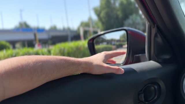 Close-up of a bare arm resting on a car door panel with hand near the side mirror while driving on a sunny urban highway with green trees and blue sky.