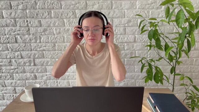 A young woman wearing glasses and a headset works hard in her home office. There is a white brick wall in the background