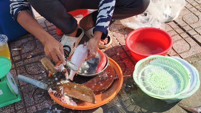 Vendor Scaling fresh fish with knife at traditional Asian street market among colorful plastic basins. Local trade features Vendor Scaling daily catch to prepare raw seafood for customer at wet market