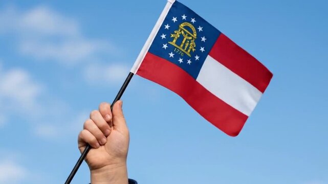 Child's hand holding the Georgia State Flag against a clear blue sky background.
