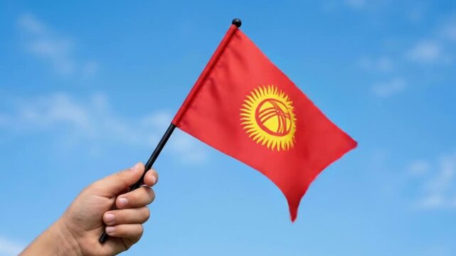 Hand Holding Small National Flag of Kyrgyzstan Against Blue Sky Background