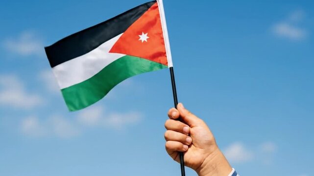 Hand Holding the National Flag of Jordan Against a Clear Blue Sky