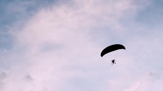 A paraglider soaring through a cloudy sky. The paraglider and canopy create a silhouette against the expansive backdrop of the sky.