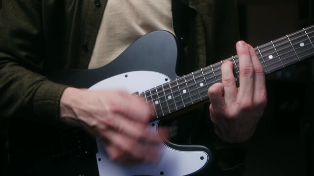 Close up guitarist hands playing rhythm on black and white electric guitar, strumming strings and fretting chords on fretboard in dark studio setting. Musician strumming, playing electric guitar riff