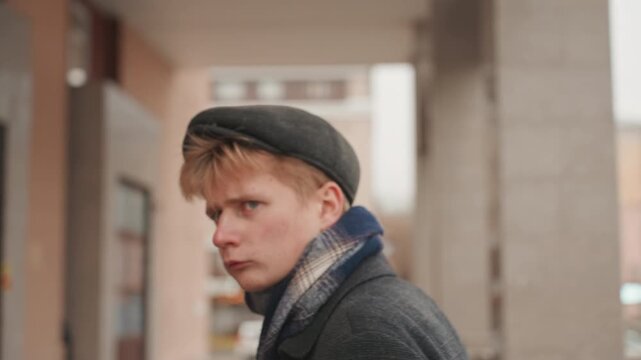 White creator scouting urban location under overcast sky, coat and cap, purposeful walk past columns and storefronts, pensive look toward street, cinematic framing for film or photo scouting