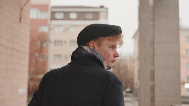 White poet creator walking through city, rear and closeup shots of young white man in wool coat, cap and scarf, sideways glances, introspective mood, muted overcast light and textured urban pillars