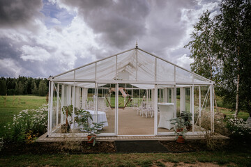 Fototapeta premium Glass greenhouse structure with white tables and chairs set up for an outdoor event, surrounded by greenery and under a cloudy sky in a rural landscape