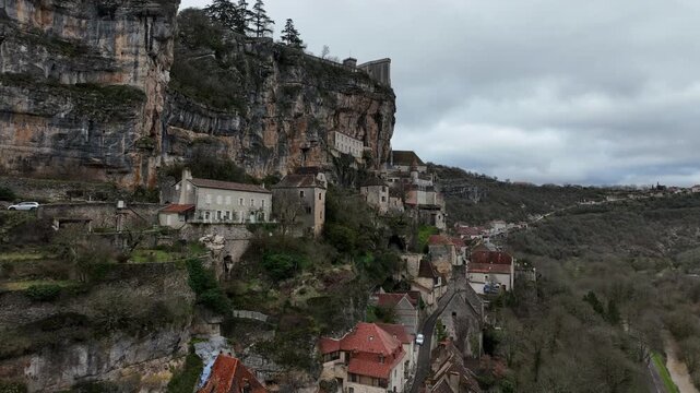 Aerial view of Rocamadour dramatic village and sanctuary buildings clinging to steep rocky cliff face above deep gorge under cloudy sky