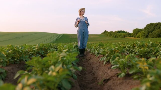 A young woman wearing overalls and a straw hat stands with her arms crossed in a vast potato field during golden hour
