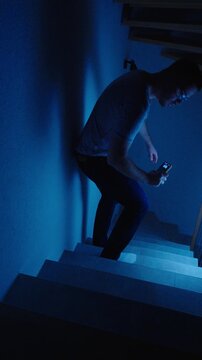 Vertical low-light shot of a frightened man walking down stairs with his smartphone during a power outage, heading to the basement fuse box and showing fear, uncertainty, and home emergency tension.
