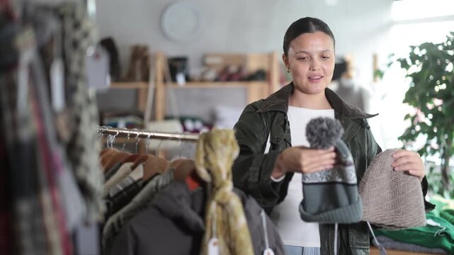 While shopping, Asian woman browsing goods items and choose elect warm hat in store, buyer consider some items. Concept of seasonal wardrobe shopping. Client and holiday shopping.