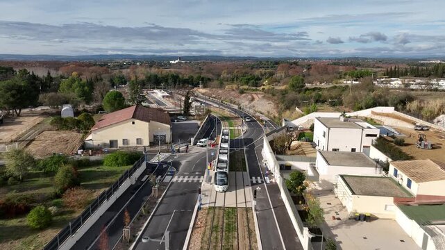 Aerial view of Montpellier tramway modern tram on elevated tracks curving through suburban area with roads buildings and green landscape under cloudy sky