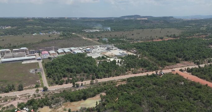 Forward aerial view revealing a concrete mixing plant with tall silos, cement mixer trucks, and large aggregate piles and industrial buildings surrounded by dense forest.