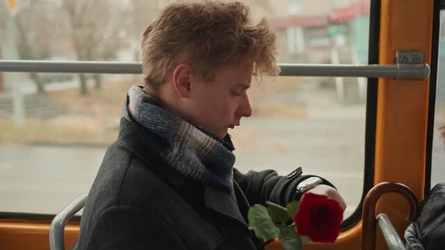 Gentle white male creator holding rose offering bloom to fellow passenger, buttoned coat and patterned scarf, window backdrop of city street, intimate gesture and hopeful romance, candid exchange