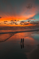 Fototapeta premium Two figures stand on a reflective beach at sunset, bathed in the dramatic hues of an orange and blue sky over the ocean, creating a picturesque and serene coastal scene.
