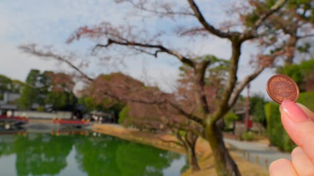 A woman's hand holds a 10 yen coin in front of Uji Temple. The camera pans to reveal the stunning Uji Temple Sanctuary and its reflection on the water on a clear winter morning in Uji, Japan. Features