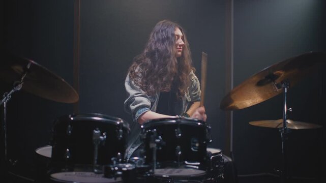Drummer girl playing drums in music rehearsal studio filled. Rock band music artist learning a drum solo. Young woman with long curly hair, fill or break on drum kit. Female drumming in room