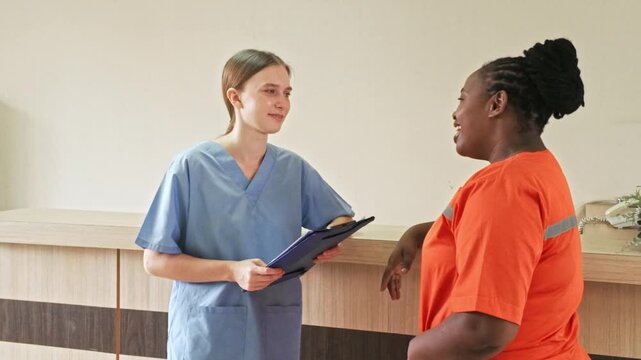 A healthcare worker in blue scrubs talks with a patient at a clinic reception, holding a clipboard during a friendly consultation.