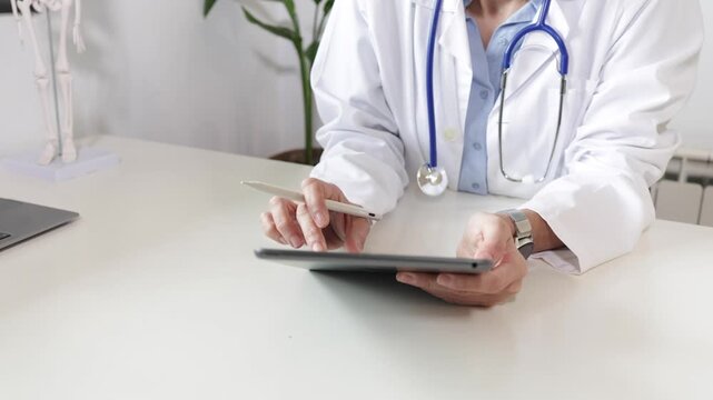 Professional female doctor in white coat using digital tablet and stylus pen for telemedicine consultation in modern clinic office, managing patient electronic health records