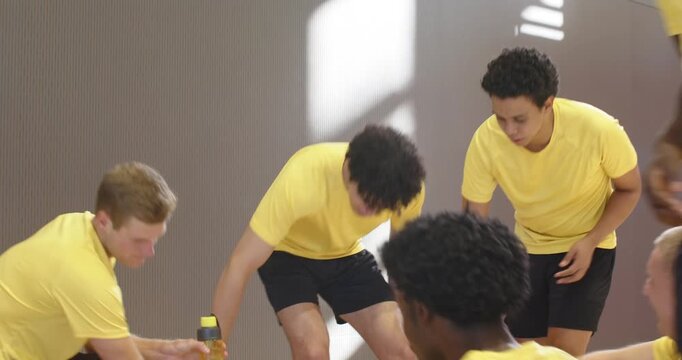 Diverse male teammates in yellow kit on bench in gym during timeout discussing play passing bottles