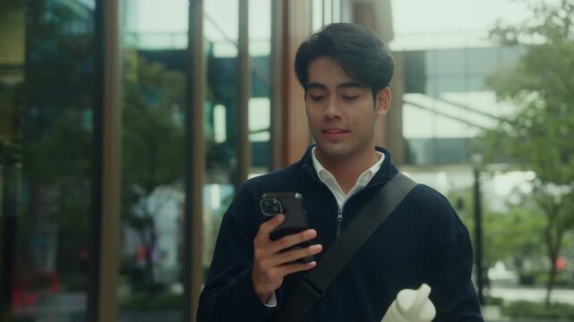 Young office worker walks through the city with a reusable water bottle in one hand, smartphone in the other, checking messages and emails, syncing calendars, and reviewing his to-do list on the go
