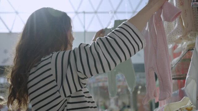 Expectant young mother carefully selecting and comparing cute baby clothing, choosing between pink and green rompers while shopping in a children's department store for her newborn