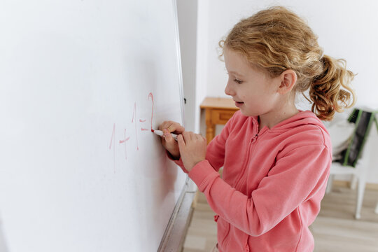 Little girl writing math problem on whiteboard indoors, active home learning moment, childhood education, homeschooling, problem solving, school routine, concentration and everyday life.
