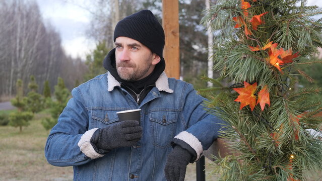 Man wearing a beanie, denim jacket, and gloves, standing outdoors holding a disposable coffee cup near pine branches decorated with autumn leaves and string lights, looking away thoughtfully