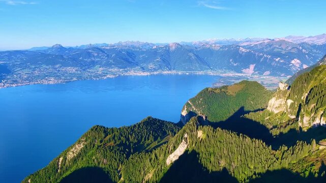 Aerial drone 4K footage featuring a smooth lateral flight along an alpine ridge overlooking Lake Leman. The shot reveals forested slopes, cliffs, and a vast mountain landscape under a clear blue sky. 